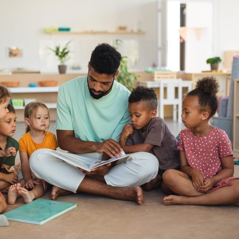 A group of small nursery school children with man teacher sitting on floor indoors in classroom, reading fairy tale book.